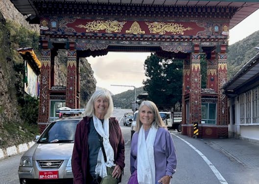 Two adult, White women stand in front of a highway gate built on the road between Paro and Thimphu in Bhutan. They are smiling and it's a sunny day.