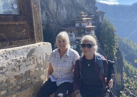 Two adult, White women, sit at the top of the hike with the famous Tiger's Nest Monastery in Bhutan in the background. It's a sunny day. They are smiling. The woman on the right is wearing black sunglasses.