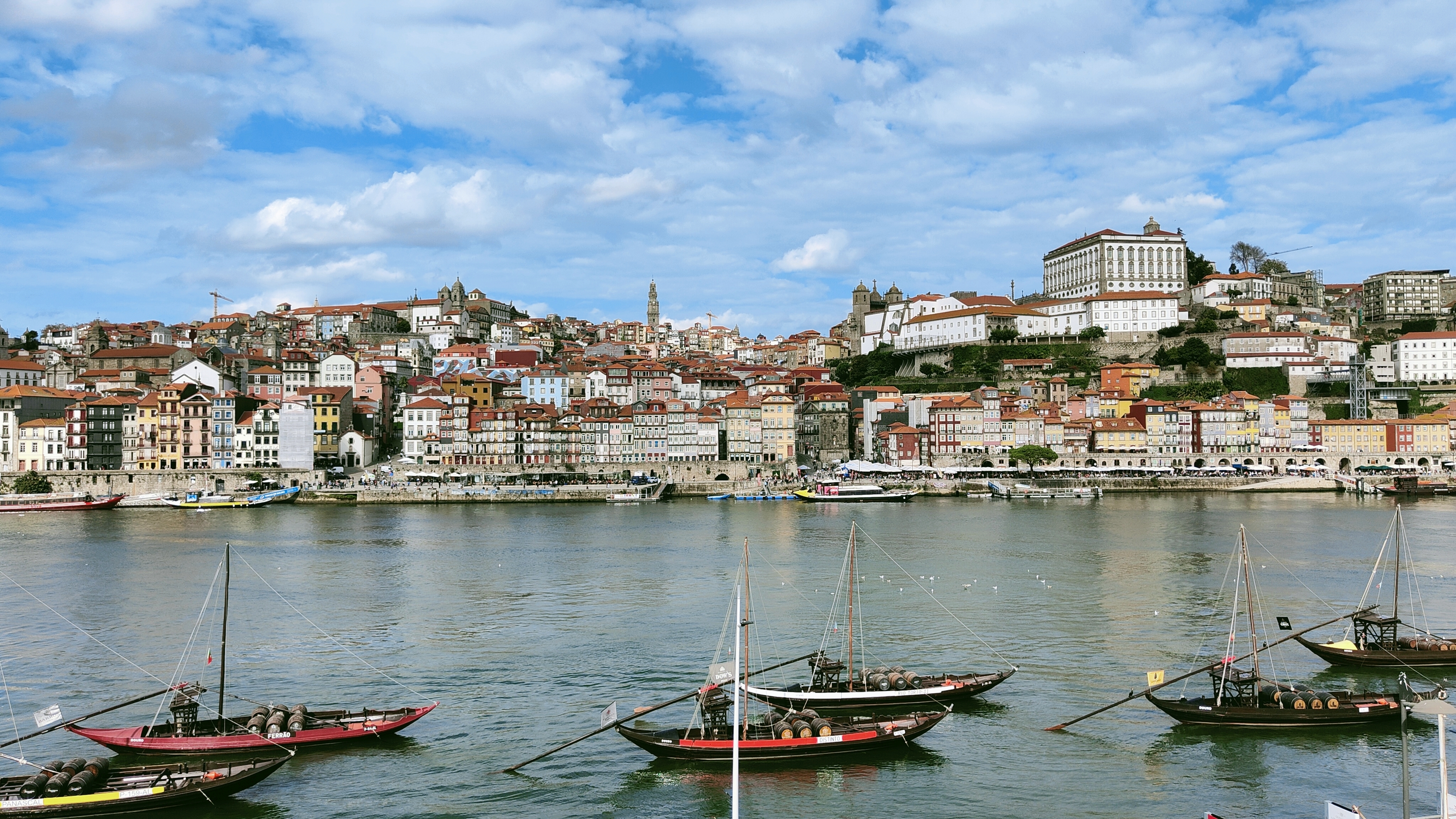 A cute, modern-looking European city with short, colorful houses line the short across a river. Small fishing and sailing boats are in the foreground. It is bright but the blue sky is mostly cloud-covered. This is in Portgual.