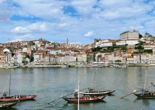 A cute, modern-looking European city with short, colorful houses line the short across a river. Small fishing and sailing boats are in the foreground. It is bright but the blue sky is mostly cloud-covered. This is in Portgual.