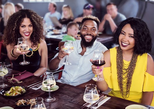 A group of three Black friends, two women with a man in the middle, face the camera and smile radiantly, each holding a glass of wine. They are in South Africa in the Winelands on a sustainable slow travel tour.