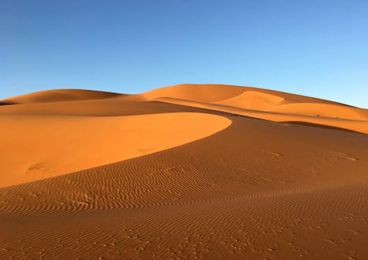 The sand dunes of Erg Chebbi in Morocco are shown in soft orange, some dunes shaded, under a blue sky.