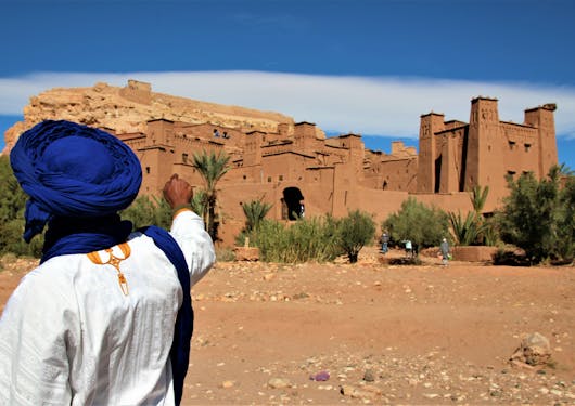 A local Berber man in white garb and a blue head wrap points at the fortified buildings of Ouarzazate in Morocco.