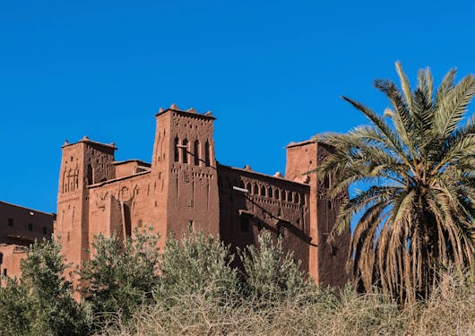 Aït Benhaddou, a historic ksar and fortified building in Morocco's Ouarzazate town, is shown with greenery in the forefront and under a bright blue sky.