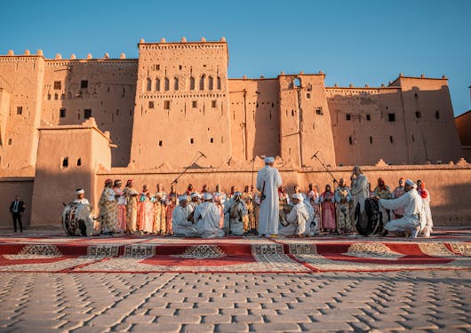 The iconic Kasbah Taourirt, a fortified buiding from the 17th century, is shown in Morocco's Ouarzazate town, with local people celebrating a festival in traditional garb in front of it on a woven rug,