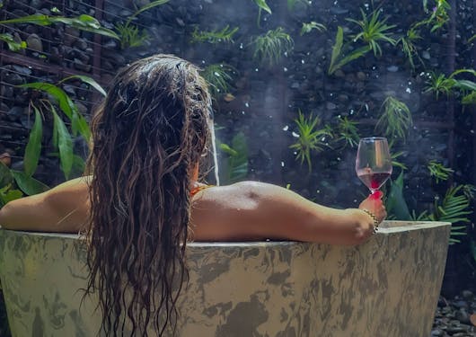 An adult woman sits in a rainforest bathtub with her back to the camera, she has long hair flowing over the side of the tub and is holding a glass of wine as she relaxes at a luxury glamping site in Costa Rica.