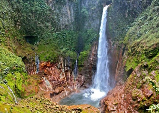 The famous Toro Waterfall is shown in Costa Rica, cascading from above into a pool surrounded by red rocks and moss-covered cliffs.