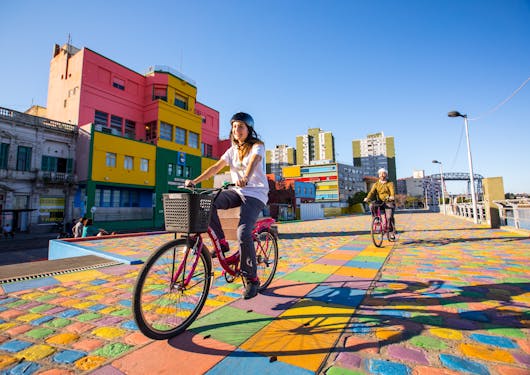 A woman rides on a bicycle on a street in Buenos Aires covered in colorful bricks with bright buildings behind her; another cyclist follows.