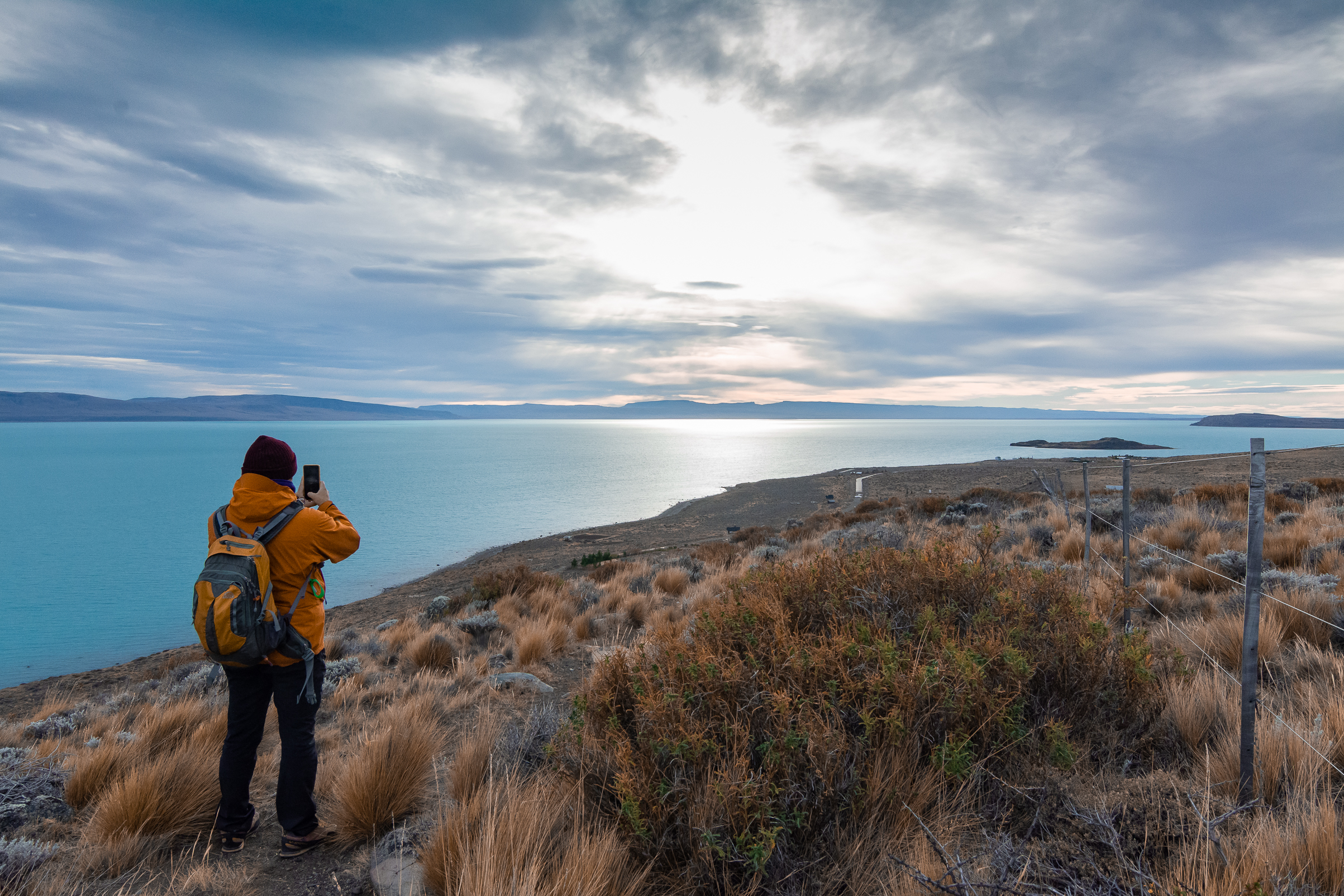 An adult bundled in a hat, orange jacket, and a backpack stands atop the cliffs in El Calafate looking over a placid blue lake in the afternoon sun.