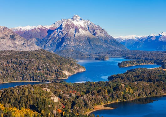 A beautiful view of Barioche, Argentina, showing its Lakes and Mountains separated by green forests under a blue sky.