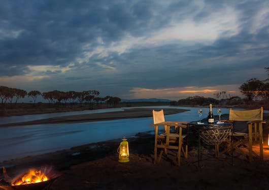 A serene camp in Kenya is shown at dusk with warm lighting along the ground.