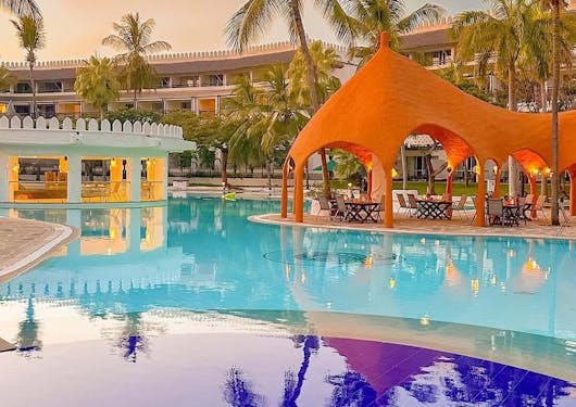 A view over a blue pool with an orange tent in the background and the royal Swahili accommodation behind, at sunset and with palm trees around.