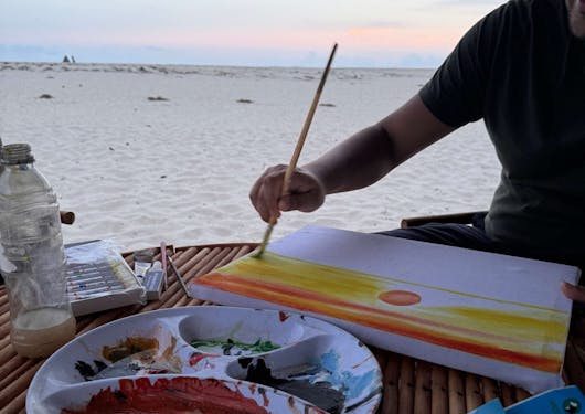 An activity of painting by the beach in Kenya is shown, as a painter dabs their brush on canvas with the ocean in the background.