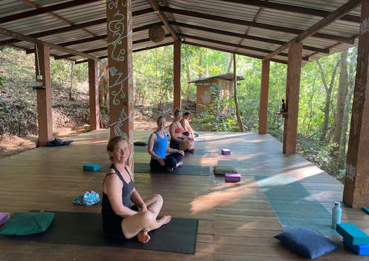 Several Caucasian women sit on their yoga mats during a yoga session in an outdoor, covered studio in Sri Lanka.