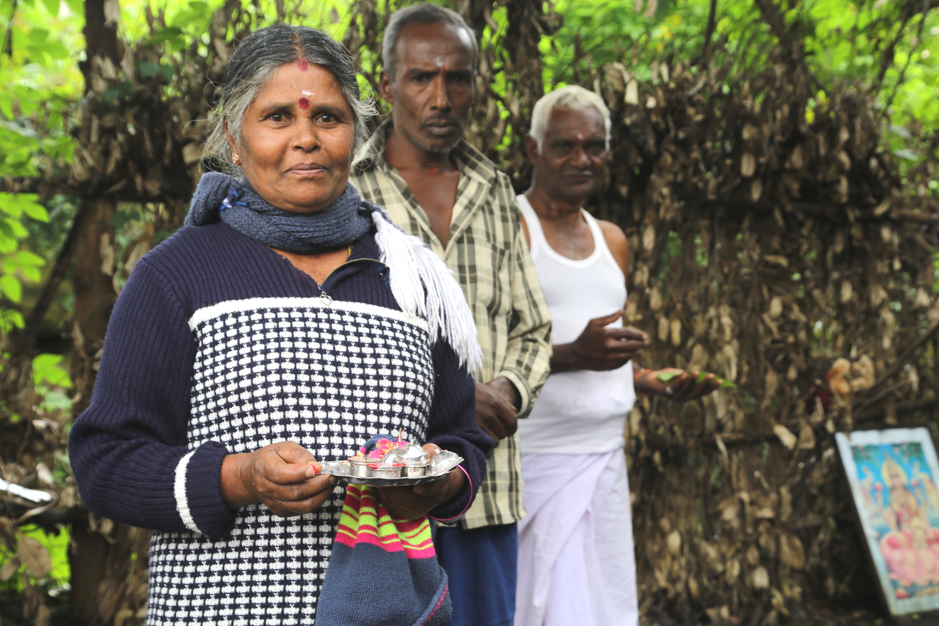 Three local Sri Lankan adults, 2 men and a woman, stand in a tea estate. The woman is holding a tray of small items.
