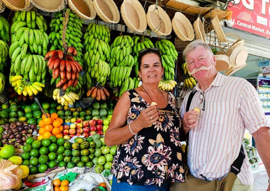 A hetero Caucasian couple stand in front of a produce market stand in Kandy with green bananas and colorful fruit behind them and wicker trays hanging overhead.