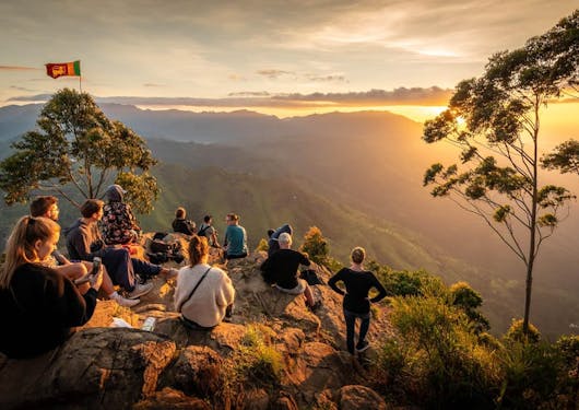 A beautiful scenic viewpoint at Ella Rock in Sri Lanka along the Pekoe Trail shows a group sitting atop a mountain watching a stunning yellow sunset.