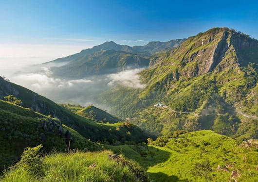 A beautiful scenic viewpoint at Ella Gap in Sri Lanka along the Pekoe Trail shows green hills with brown mountain peaks under a blue sky.