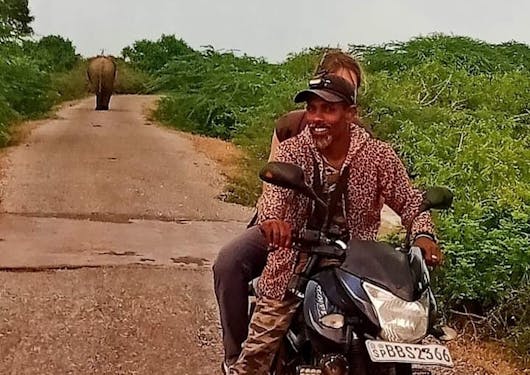 A traveler sits behind an expert local guide on a motor bike during a safari, with an elephant in the distance.