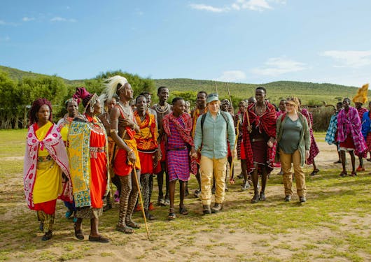 Local Kenyan Maasai Mara people stand in colorful dress with two white tourists.