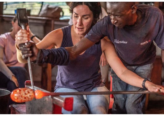 A white, adult female traveler stands over a blown glass project with a local Kenyan man.