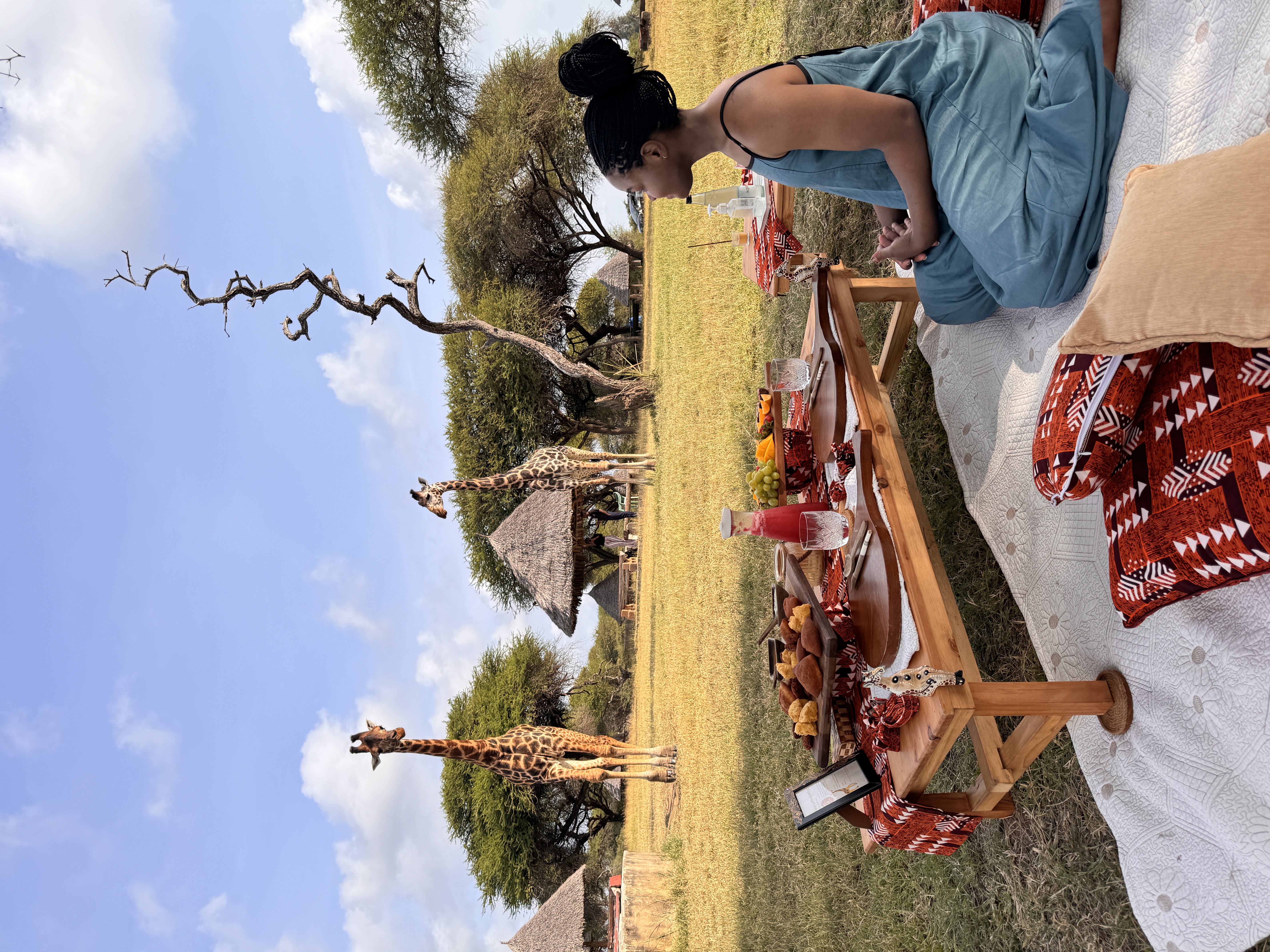 A young woman sits on a blanket in front of a picnic spread in Kenya, with giraffes in the background.