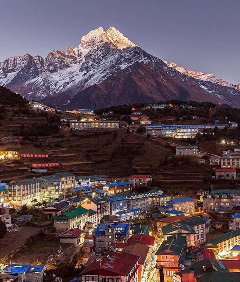 A town lit up at night is shown in front of a towering mountain in the Himalayas.
