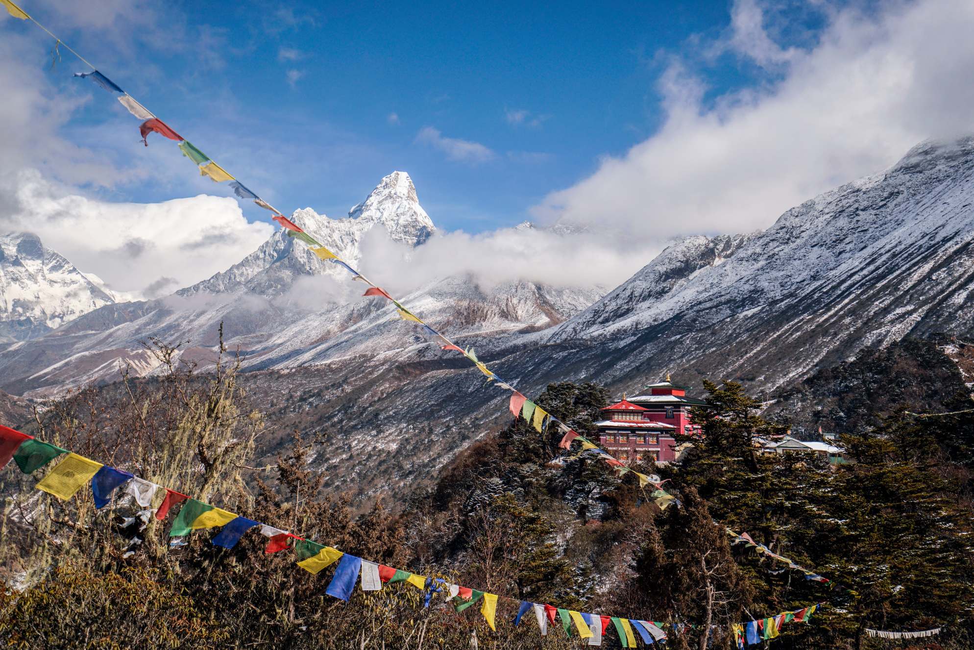 The red Tengboche Monastery is shown in the distance, with colorful prayer flags in the foreground and Mount Everest in the background.