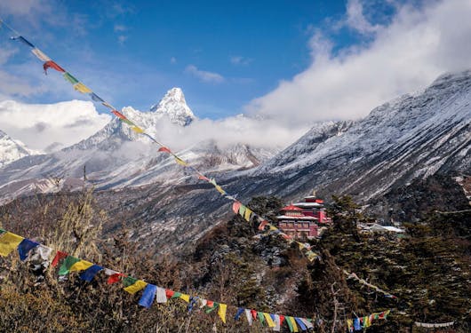 The red Tengboche Monastery is shown in the distance, with colorful prayer flags in the foreground and Mount Everest in the background.