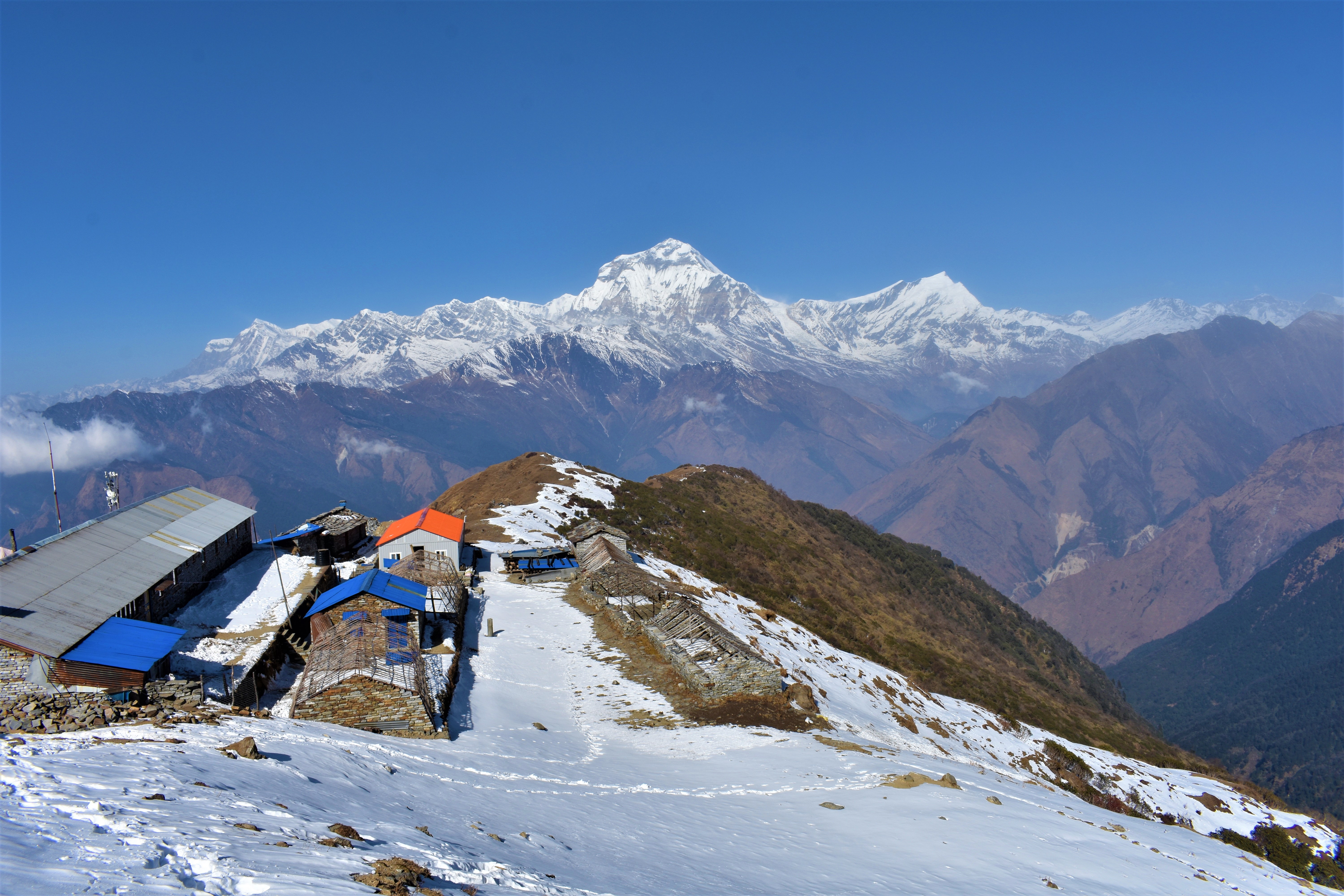 A snowy clearing with some small buildings on it is perched high and overlooking the mountains.