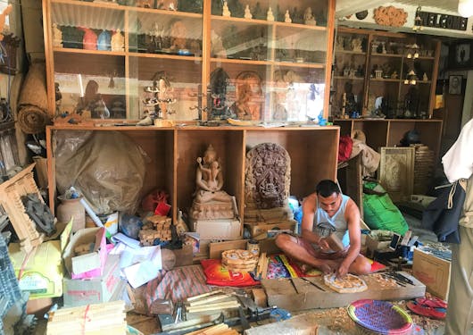 A woodcarver in Bungamati sits on the floor of his shop and works on a piece with shelves of wood products and supplies surrounding him.