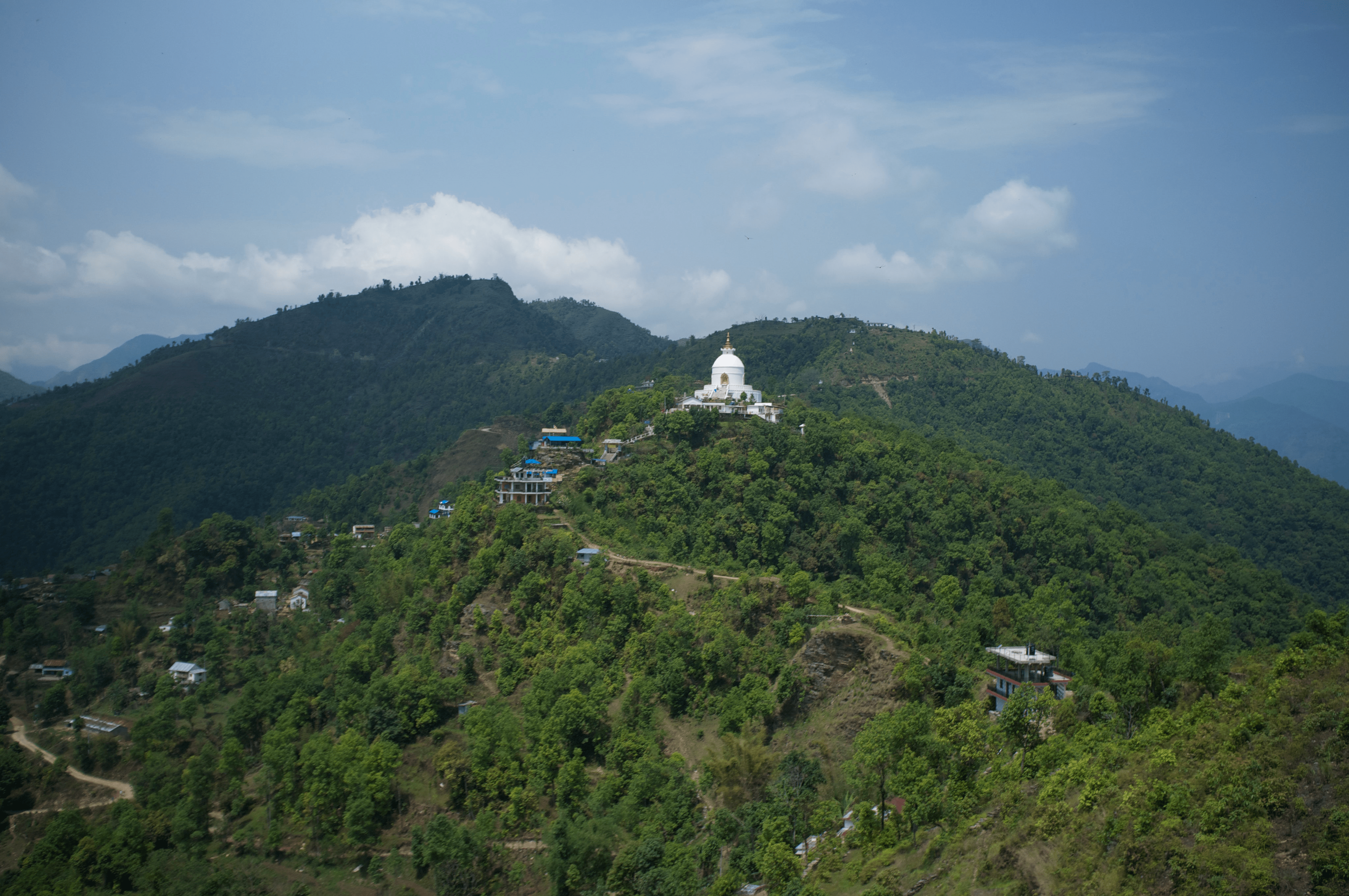 The Peace Pagoda in Pokhara is shown from a distance, a white peak perched atop a green tree-covered hill.