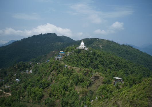 The Peace Pagoda in Pokhara is shown from a distance, a white peak perched atop a green tree-covered hill.