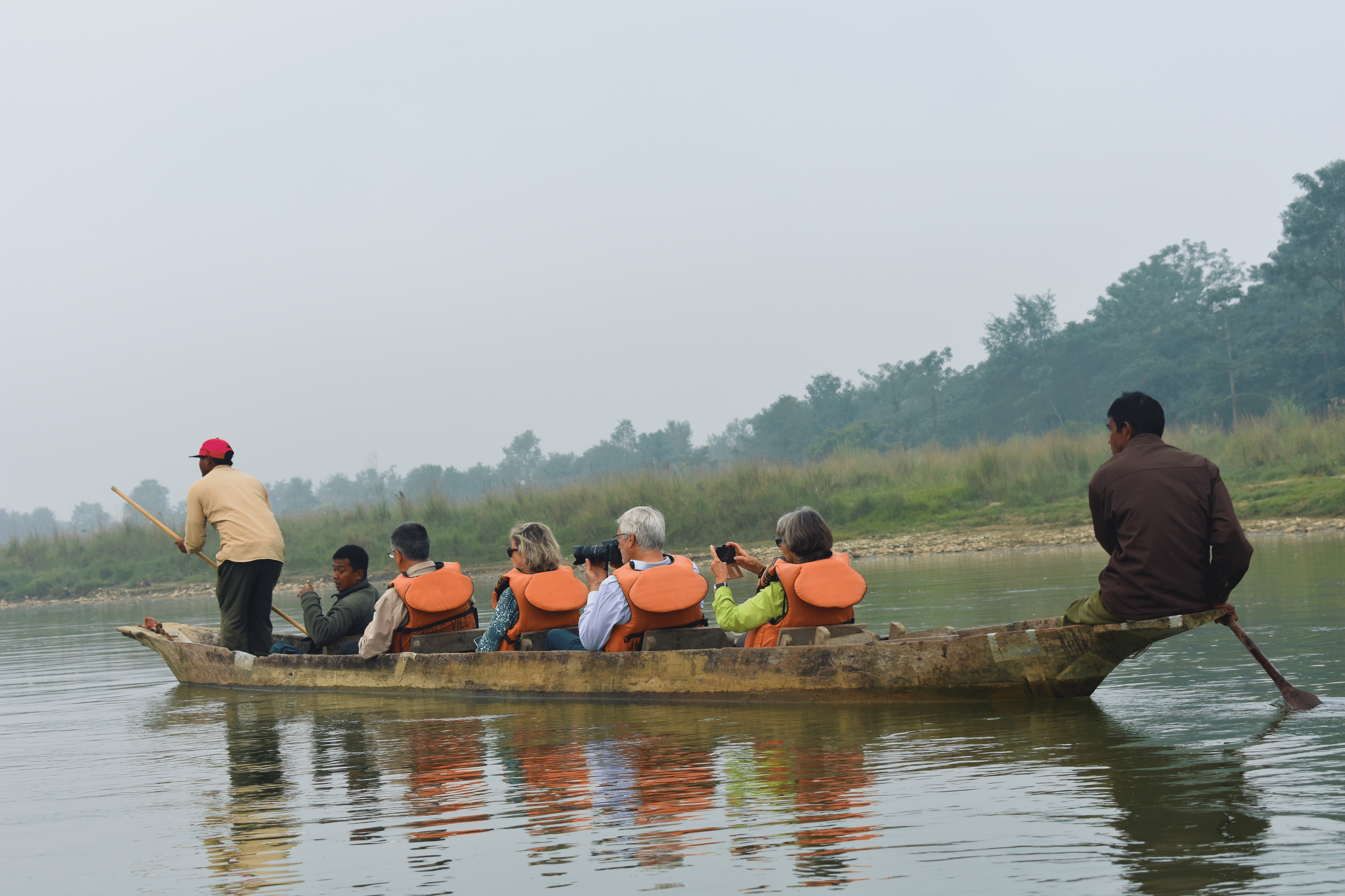 A group of travelers in orange life vests ride in a canoe paddled by two local Nepali men.