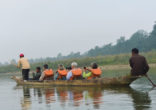 A group of travelers in orange life vests ride in a canoe paddled by two local Nepali men.