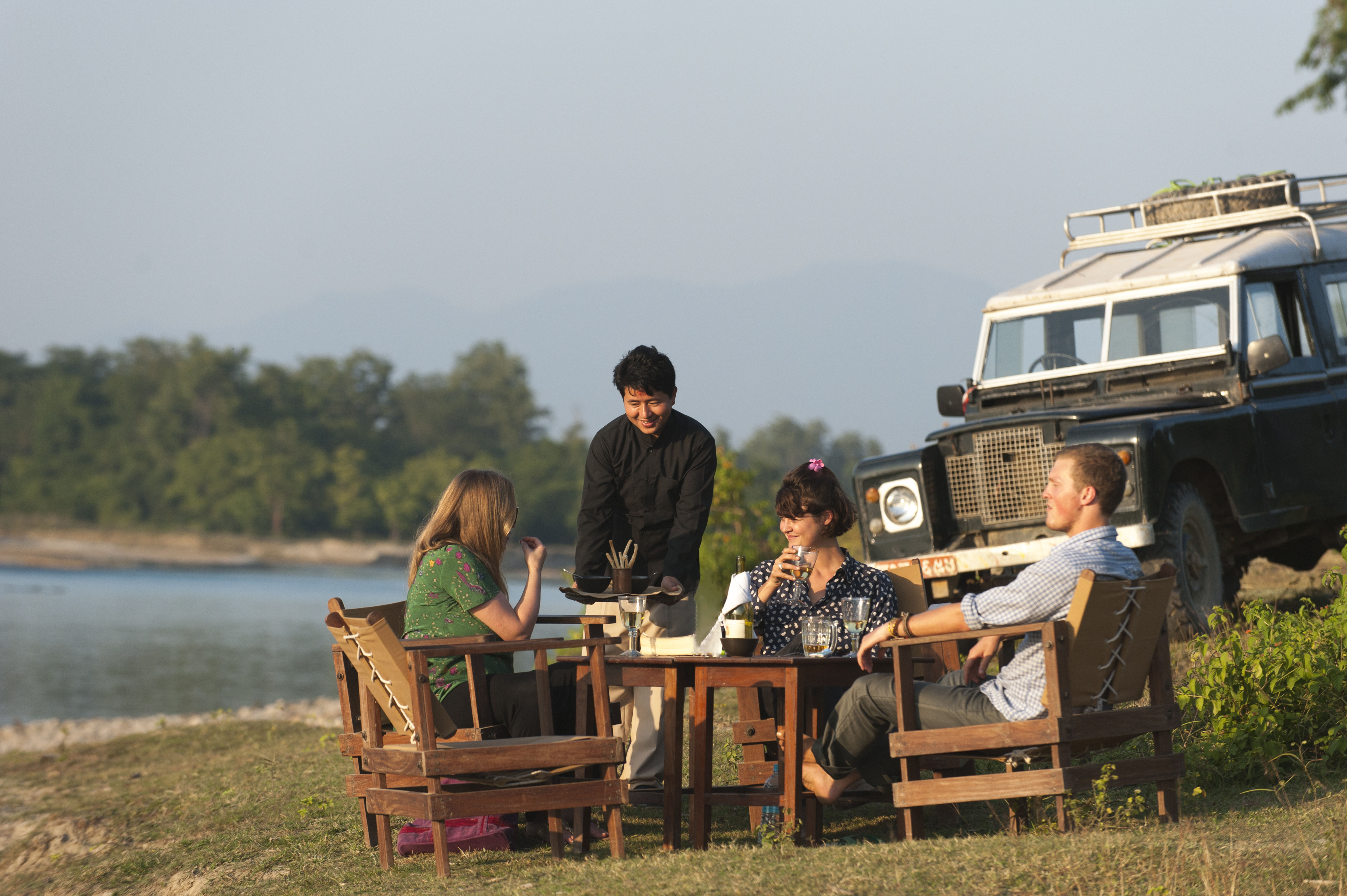 A group of 3 travelers and their local Nepali guide have dinner at sundown by a river in front of their safari jeep.