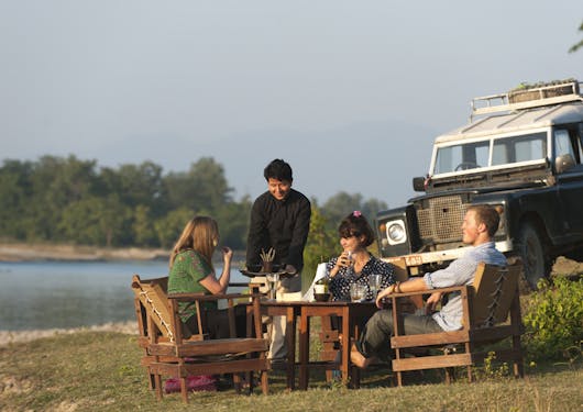A group of 3 travelers and their local Nepali guide have dinner at sundown by a river in front of their safari jeep.