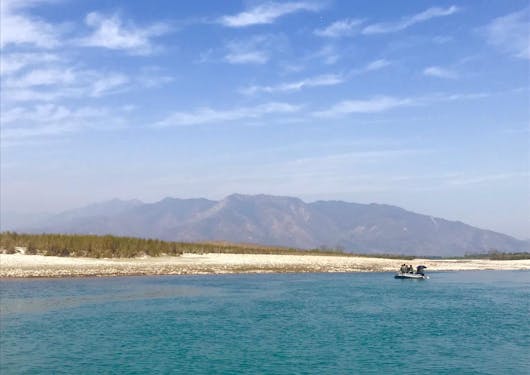 Travelers ride a river raft in the distance, down a wide, blue river in Nepal.