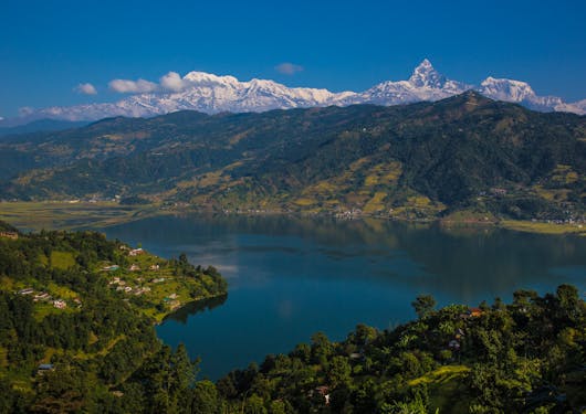 The large lake in Pokhara is shown in Nepal, surrounded by forest in all directions and with mountains in the background.