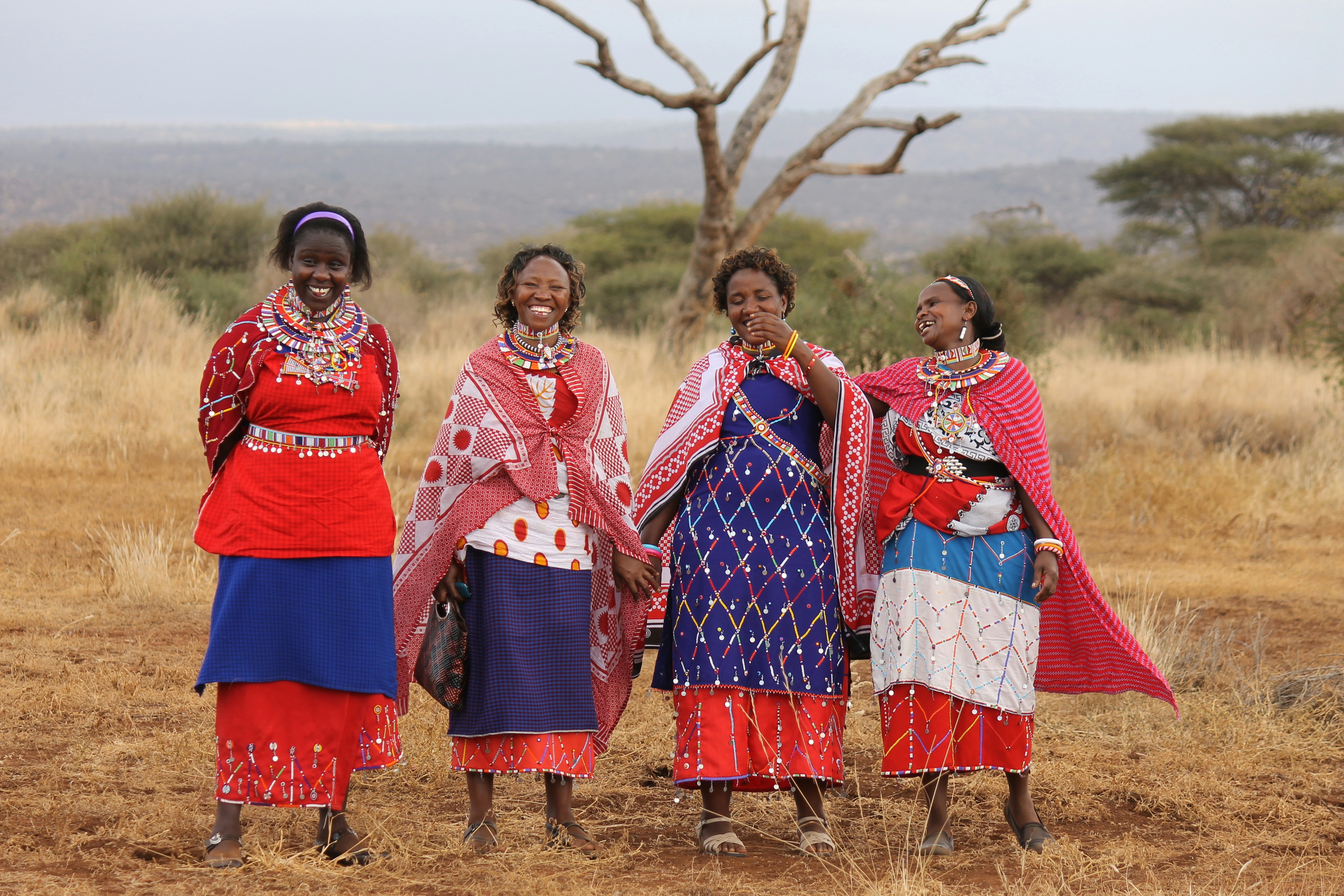 A group of four Maasai Mara women smile and pose for a photo wearing traditional attire.