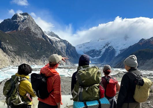 A group of 5 trekkers stand on a sunny ridge and look out over a mountain peak.