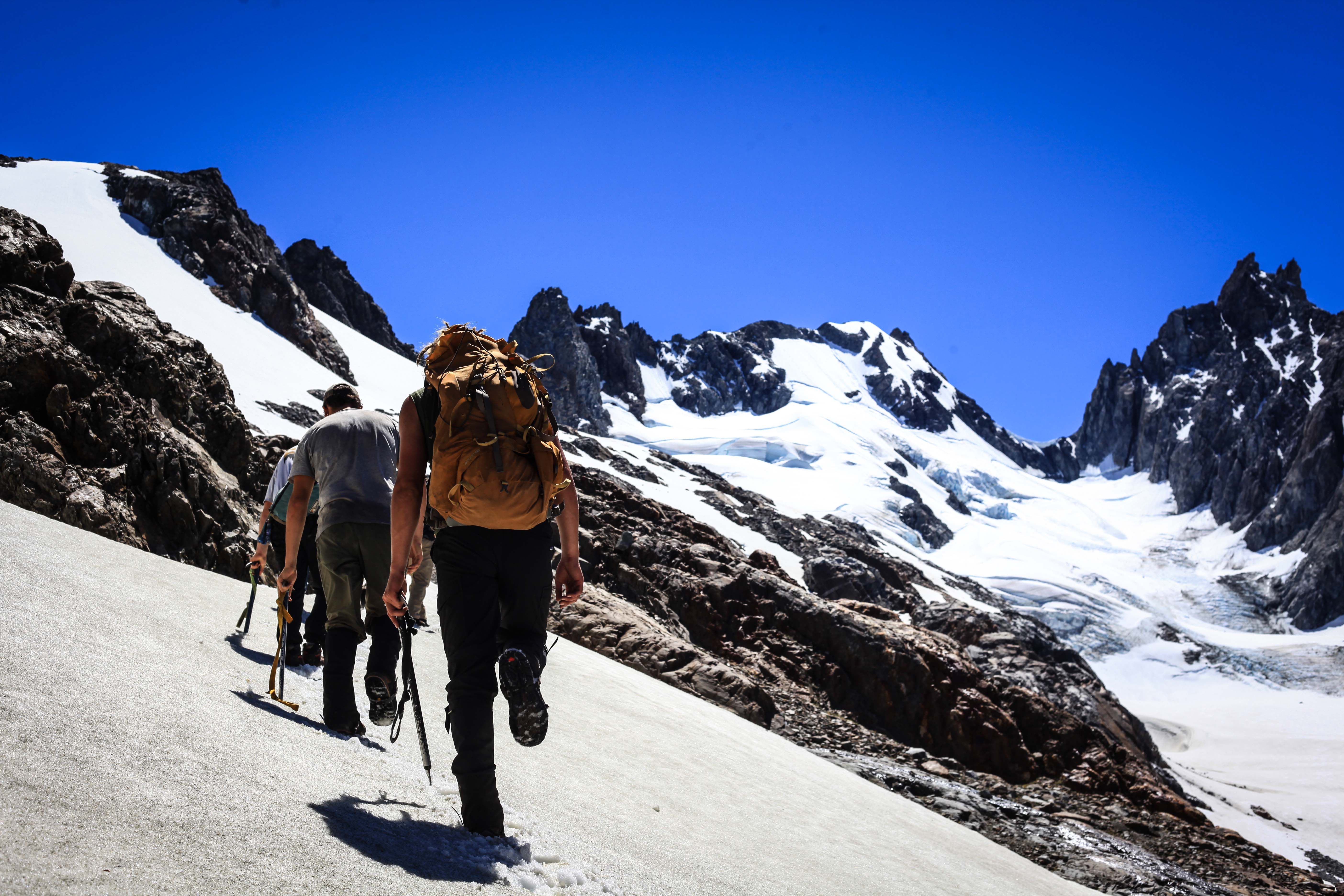 A group of trekkers hikes uphill on packed snow during a sunny day.