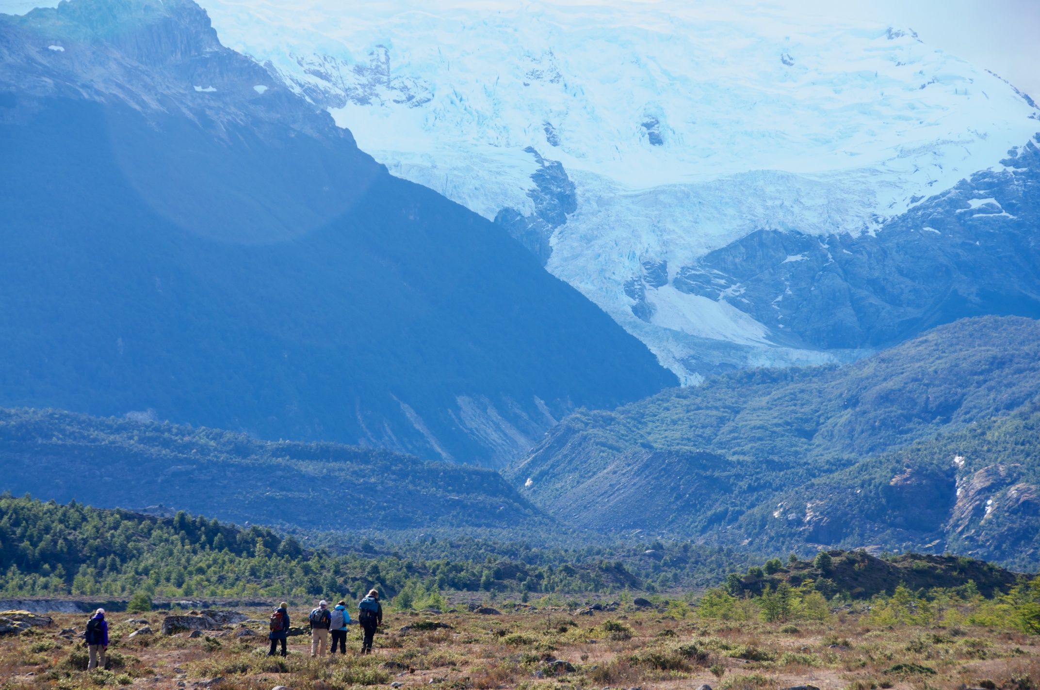 Trekkers make their way through a valley with imposing mountains in the background.
