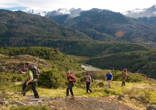 Trekkers gross a green mountain field with rolling hills and peaks behind them.