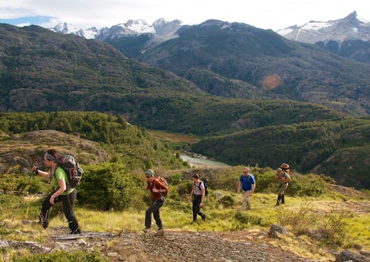 Trekkers gross a green mountain field with rolling hills and peaks behind them.