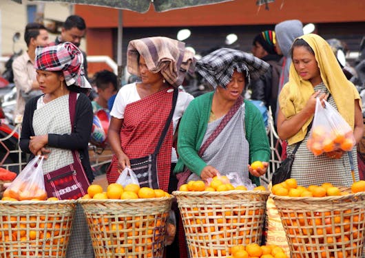 Four Indian women with head coverings stand behind citrus baskets at a market.