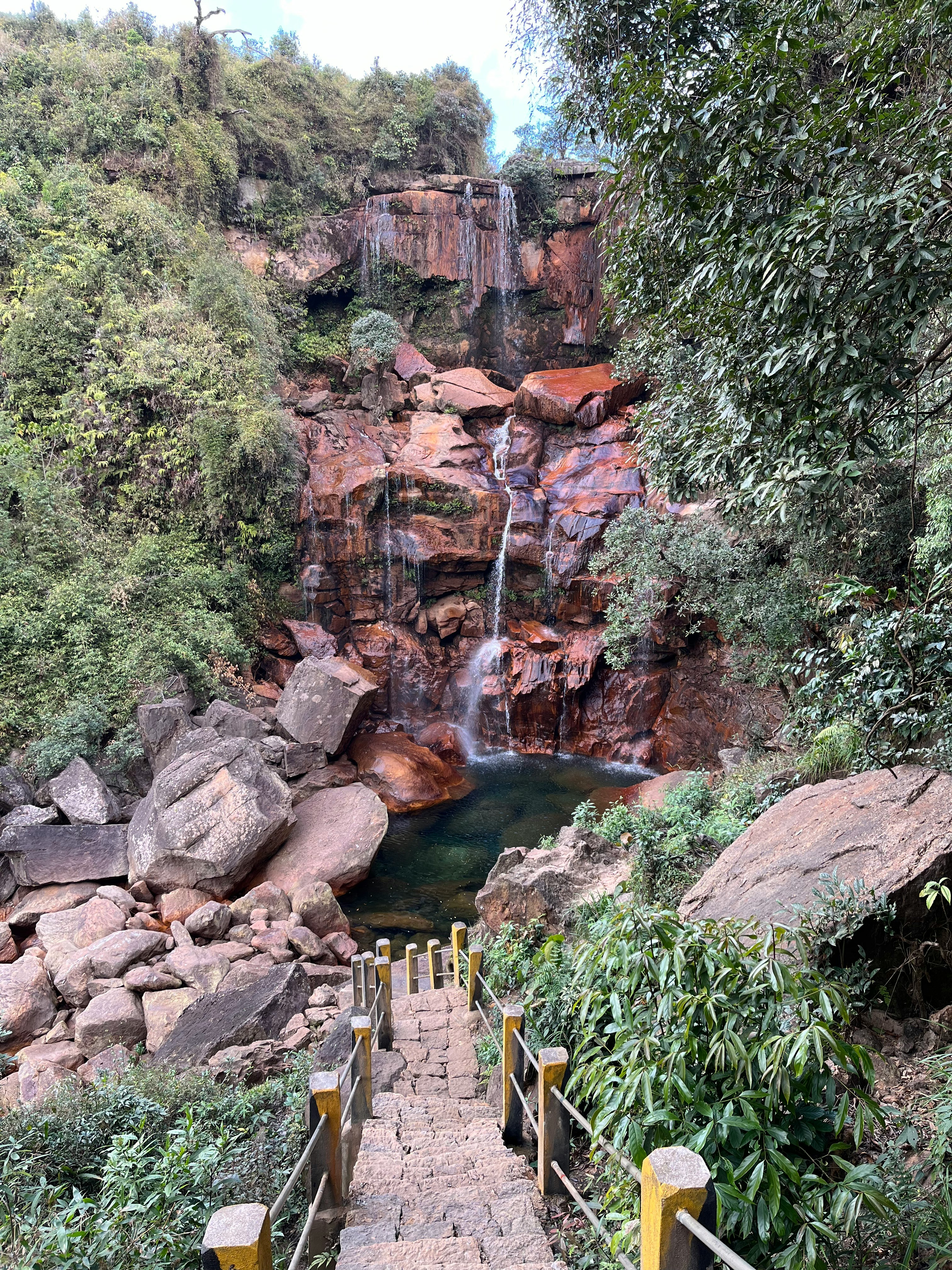 A waterfall cascading over reddish rocks with a walkway leading up to it, shown in Cherrapunjee, Meghalaya, India.