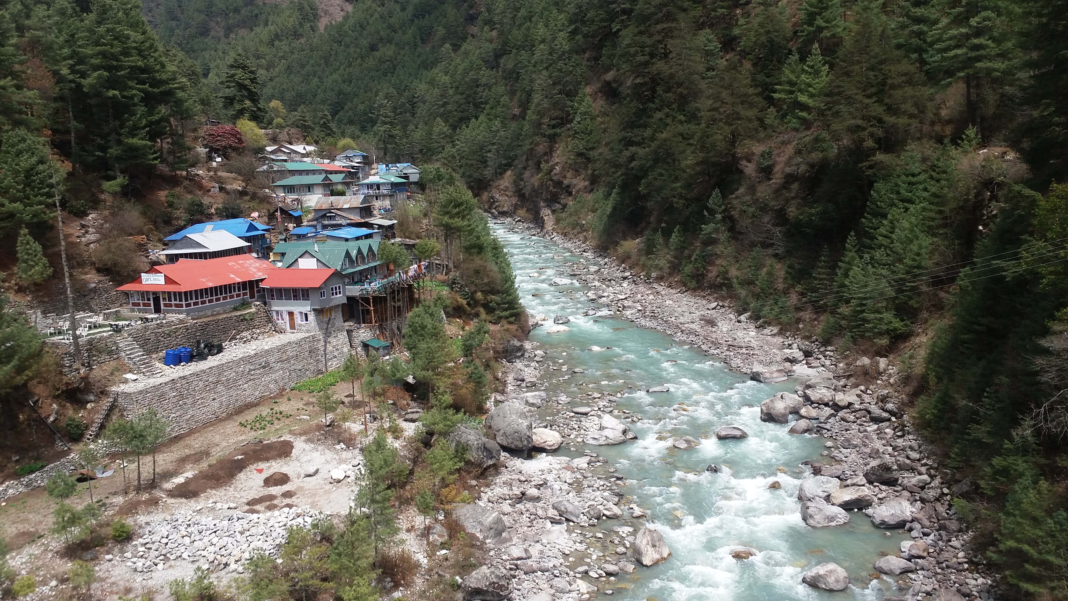 A rocky river passes nearby colorful houses as it goes through a dense green forest in Nepal.