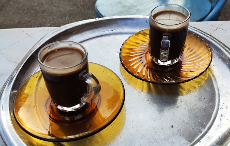 Two glass coffee cups filled with coffee site on yellow glass plates on top of a circular aluminum tray, resting on a table top with blue chairs in the background. Coffee is in in Egypt.
