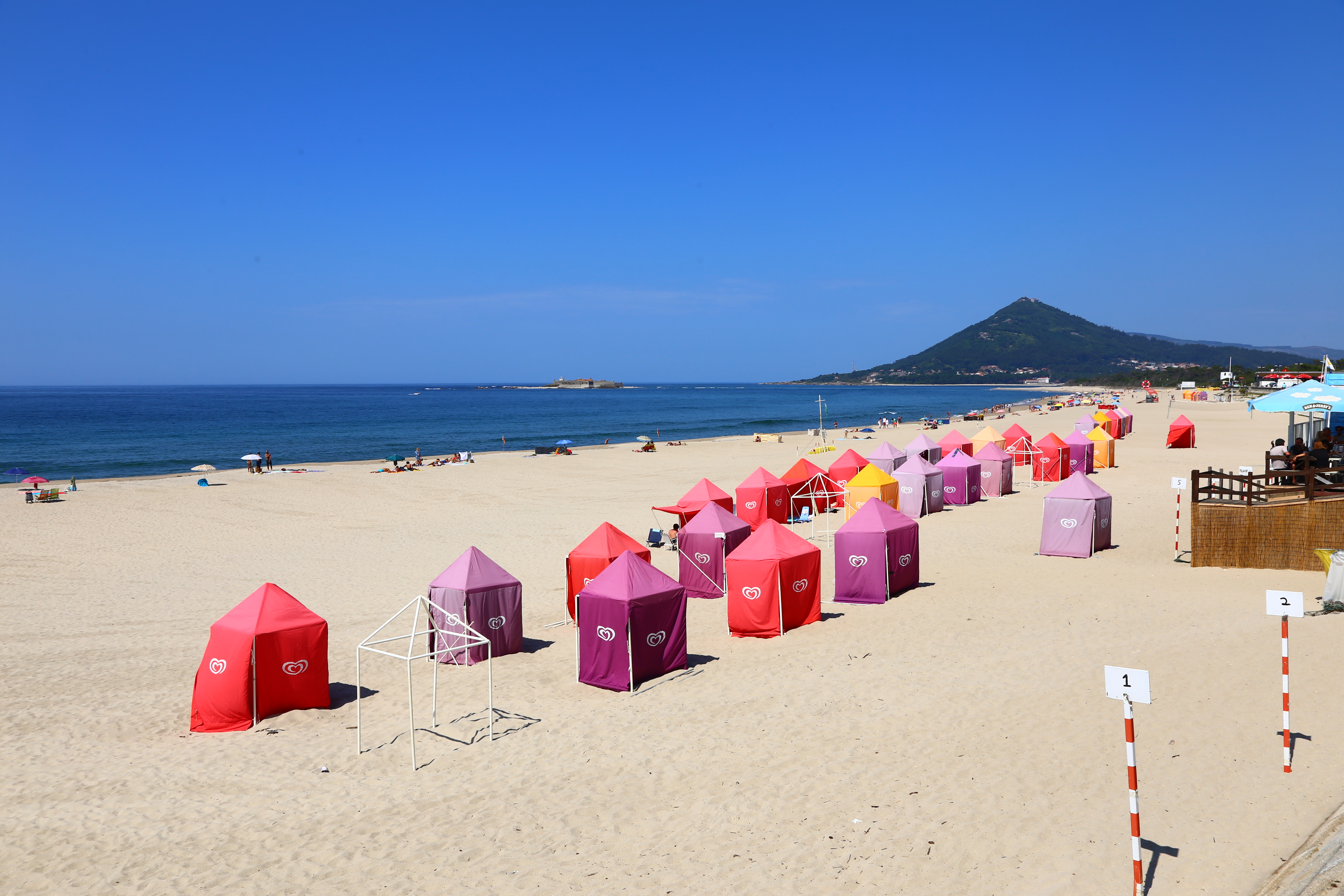 Colorful red, purple, and yellow beach huts form 3 rows on a white sane beach, with blue ocean to the left and blue sky above. This is on Portugal's Atlantic coast.